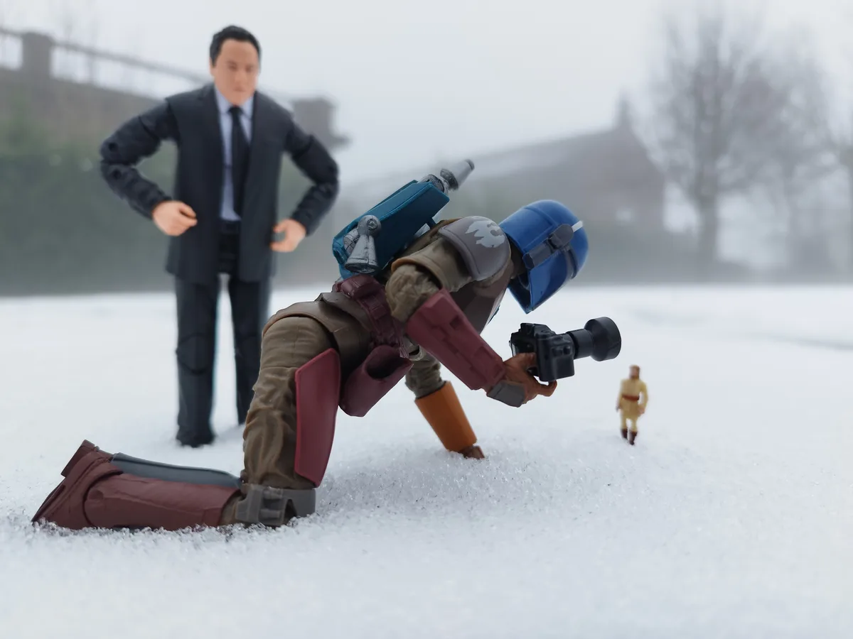 A Star Wars action figure of a Mandalorian with a blue helmet and red armor is kneeling in snow, holding a camera and…