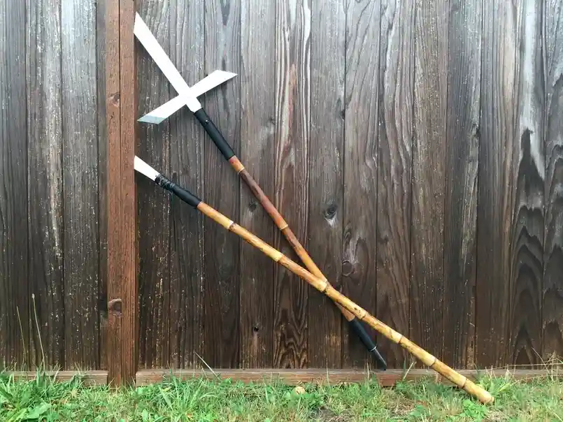 Two wooden martial arts weapons, a sword with a crossguard and a staff, lean against a weathered wooden fence. The overlaid…