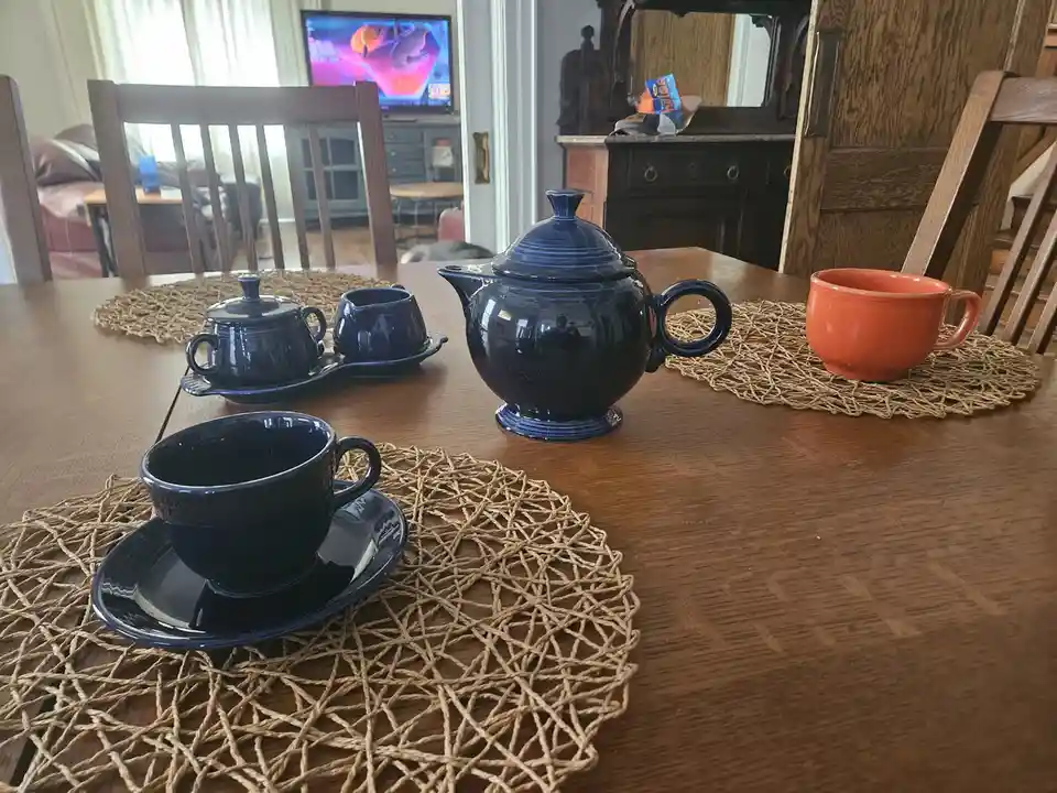 A wooden dining table set with matching blue teaware including a teapot, sugar bowl, creamer, and cups on woven placemats…