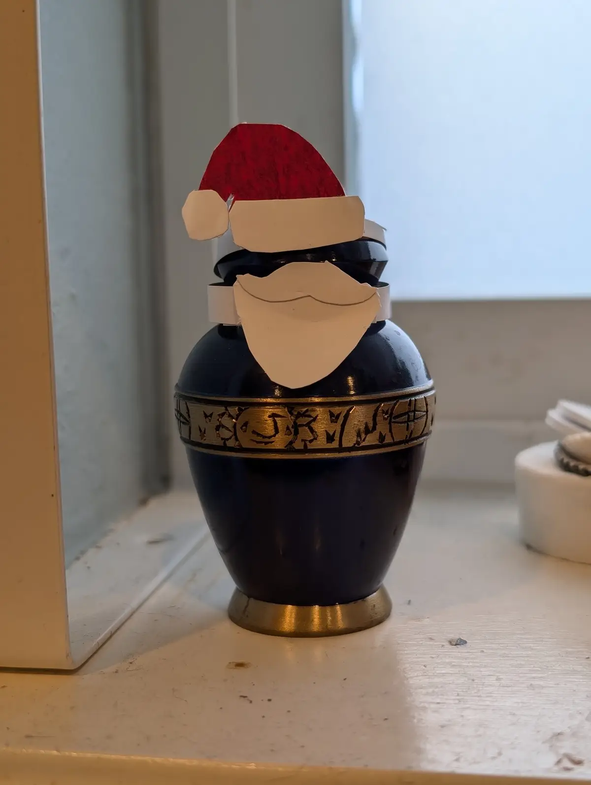 A dark blue urn with gold trim, decorated with a red paper Santa hat and white paper beard and mustache, sits on a…