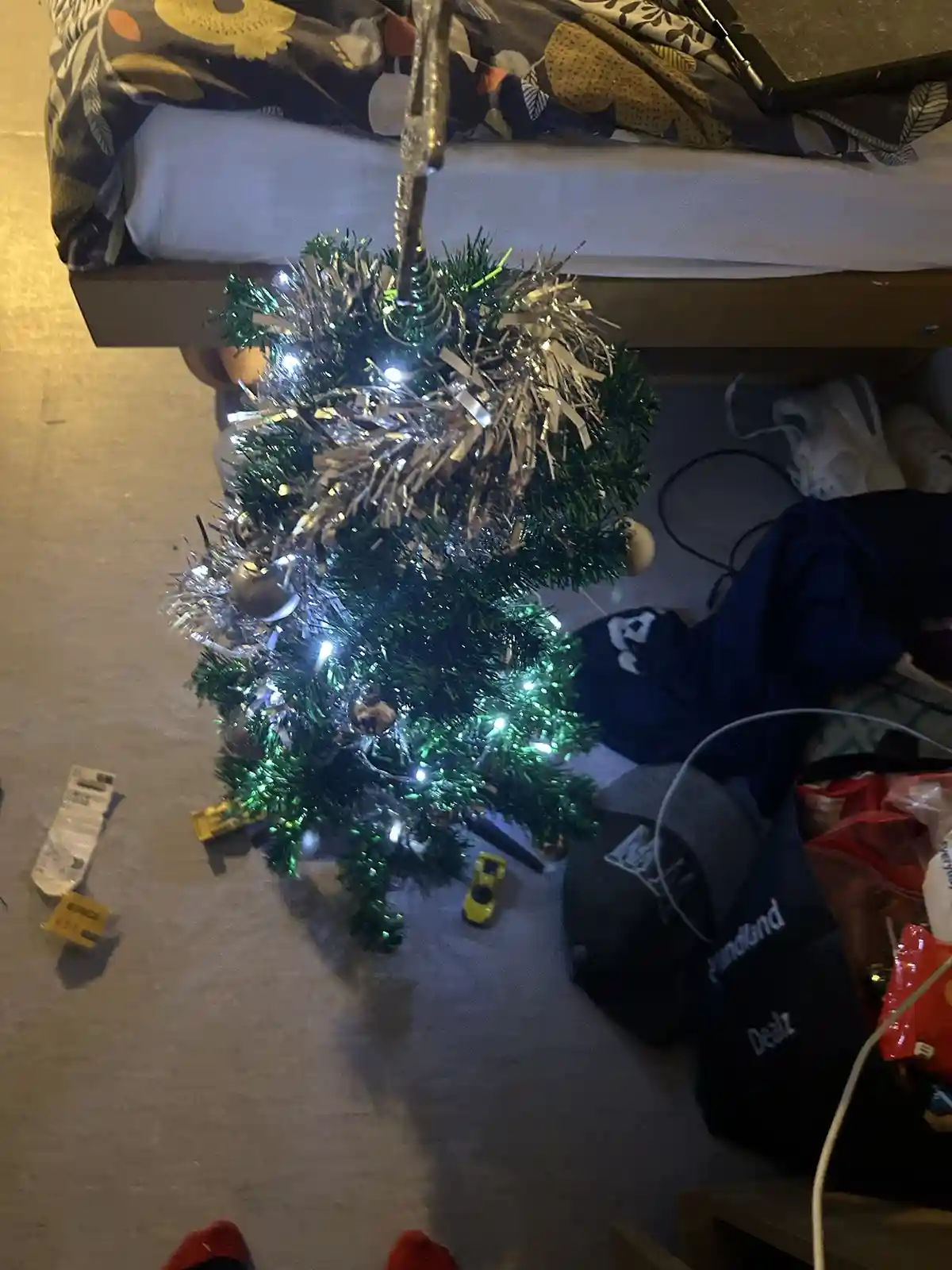 A small, decorated artificial Christmas tree with lights and tinsel is standing on a carpeted floor in a messy room, with a…