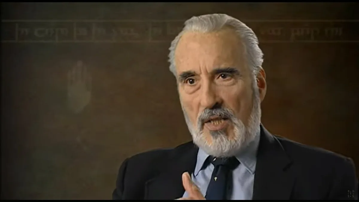 A man with white hair and a beard, wearing a dark suit and tie, is speaking with an intense expression while gesturing with…