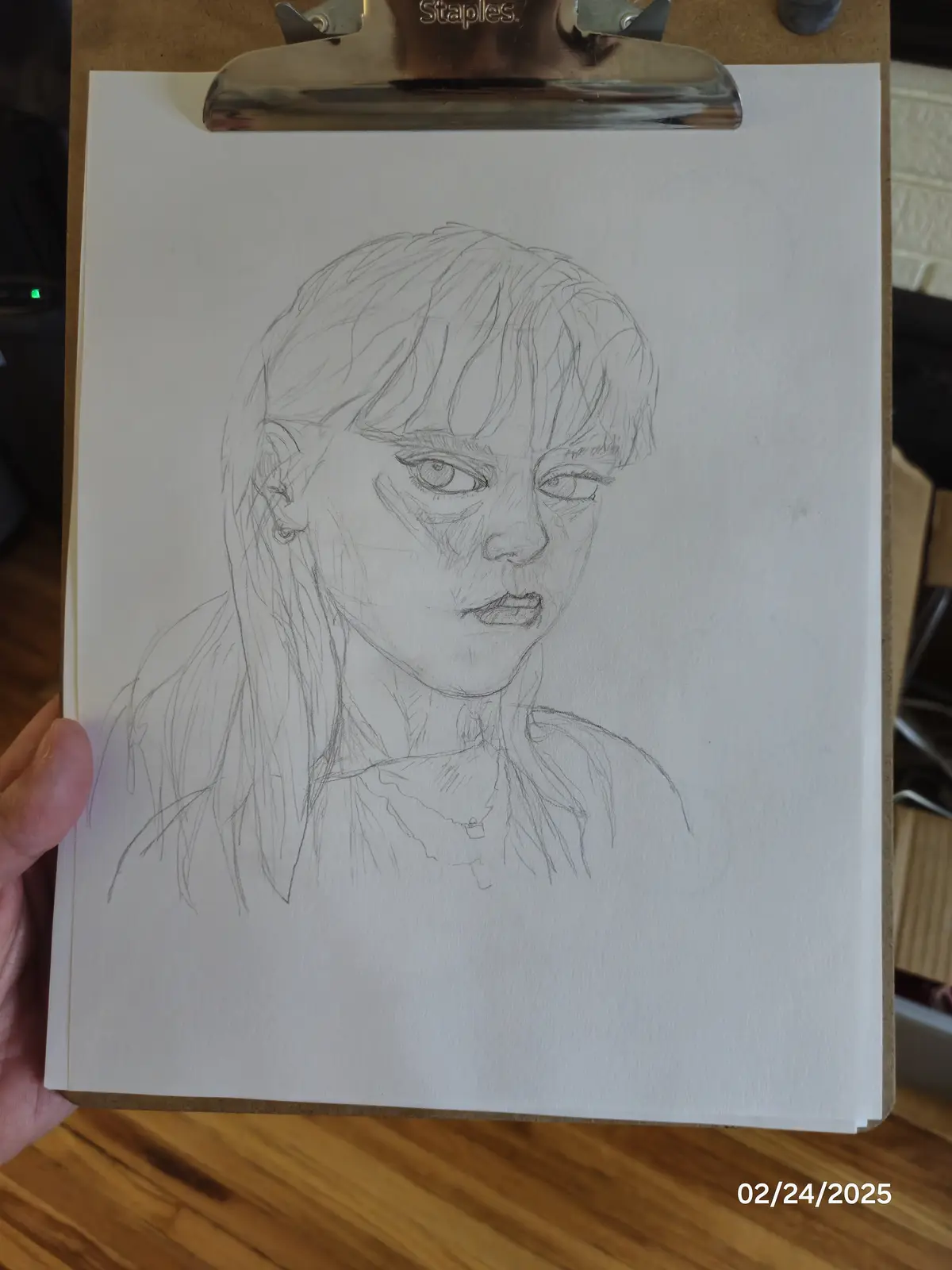 A hand holds a clipboard with a pencil sketch of a young woman's face, showing detailed shading and expressive eyes. The…