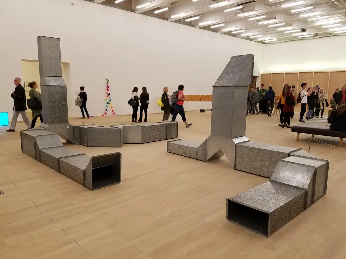 The image shows a large, abstract sculpture made of galvanized metal ductwork arranged in a complex, non-functional shape…