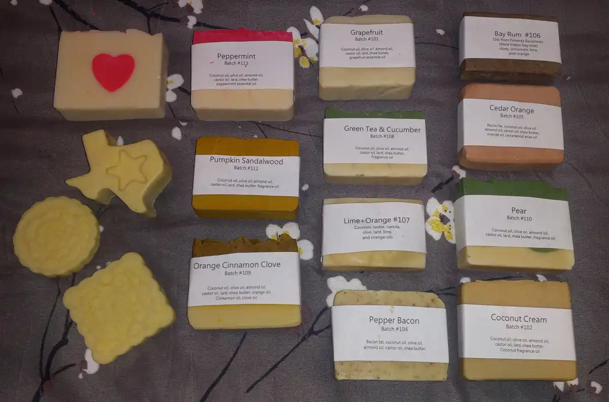An overhead shot displays a collection of handmade soap bars arranged on a floral-patterned fabric. Most are rectangular…