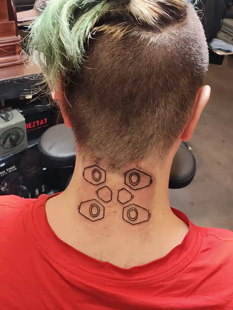The back of a person's head and neck shows a freshly outlined tattoo design of four hexagonal nuts and four hexagonal bolts…