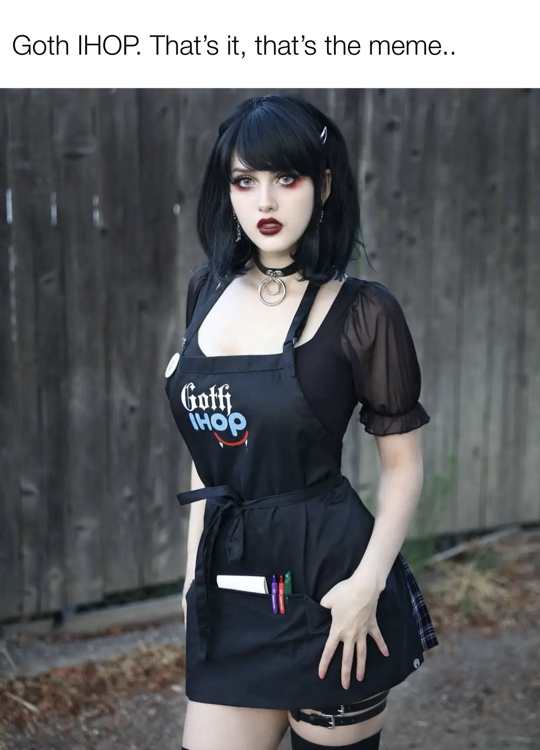 A person with goth aesthetics—black hair, dark makeup, choker, and thigh-highs—is wearing a black apron that reads 'Goth…