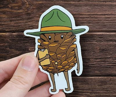 A hand holds a die-cut sticker of an anthropomorphic pinecone wearing a green ranger-style hat with a yellow band, holding a…