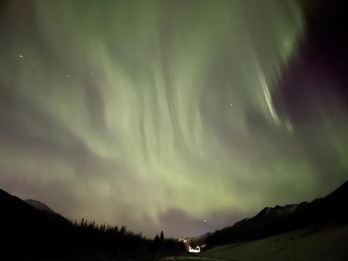 A wide-angle night photo shows vibrant green auroras dancing across a starry sky above dark mountain silhouettes and a…
