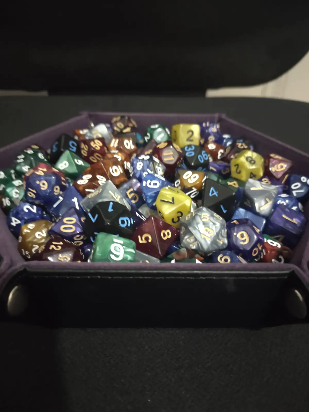 A purple hexagonal dice tray filled with numerous colorful polyhedral dice of various shapes and sizes, showing different…