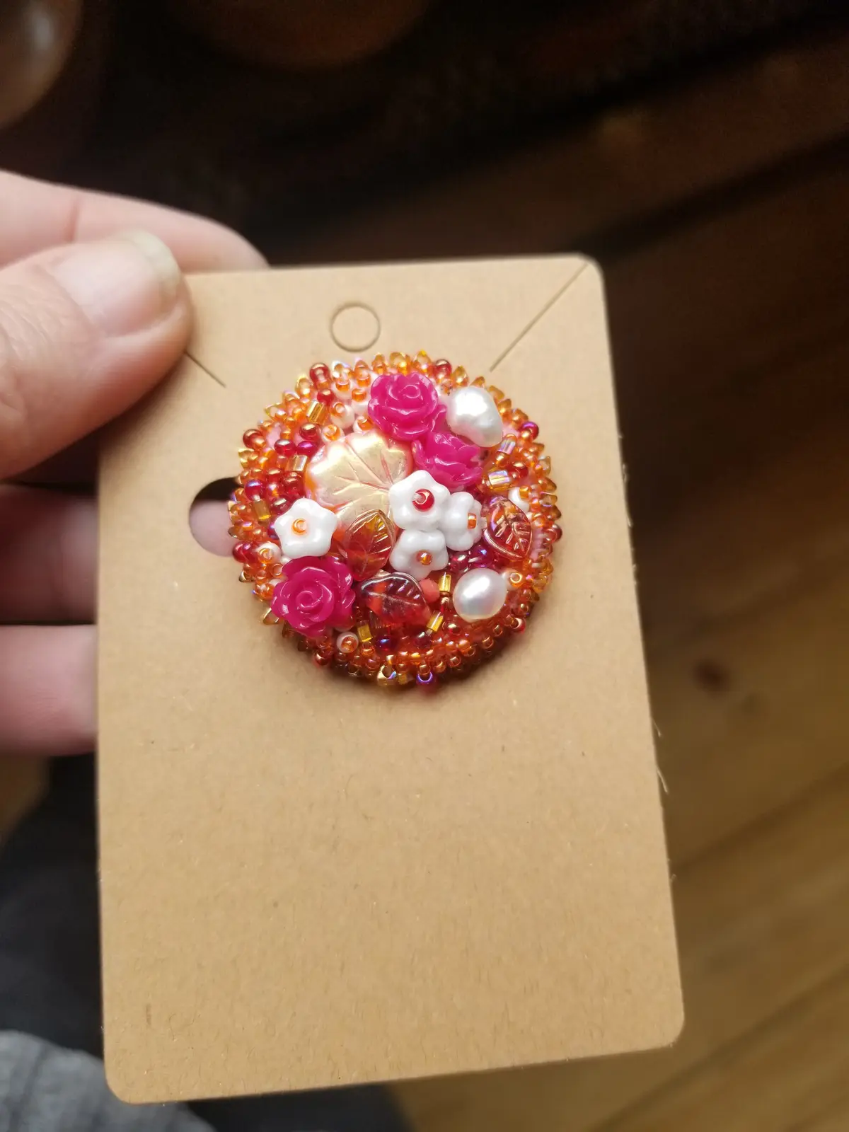 A hand holds a brown cardboard display card featuring a circular, intricate beaded brooch. The brooch is composed of…
