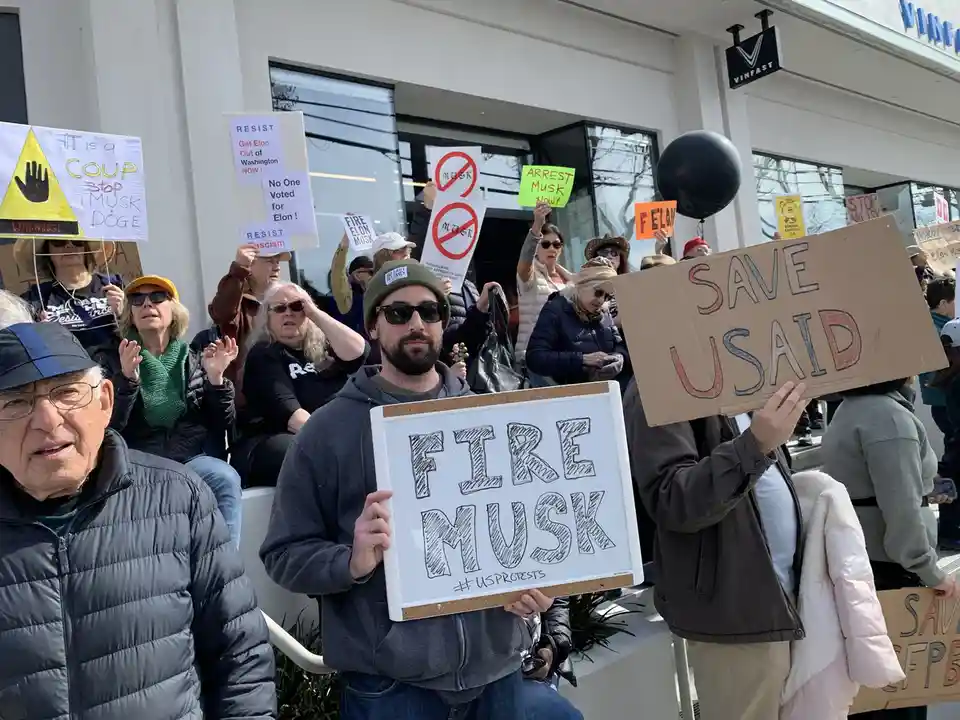 A crowd of protesters holds various signs, including one prominently displaying 'FIRE MUSK' on a whiteboard. Other signs…