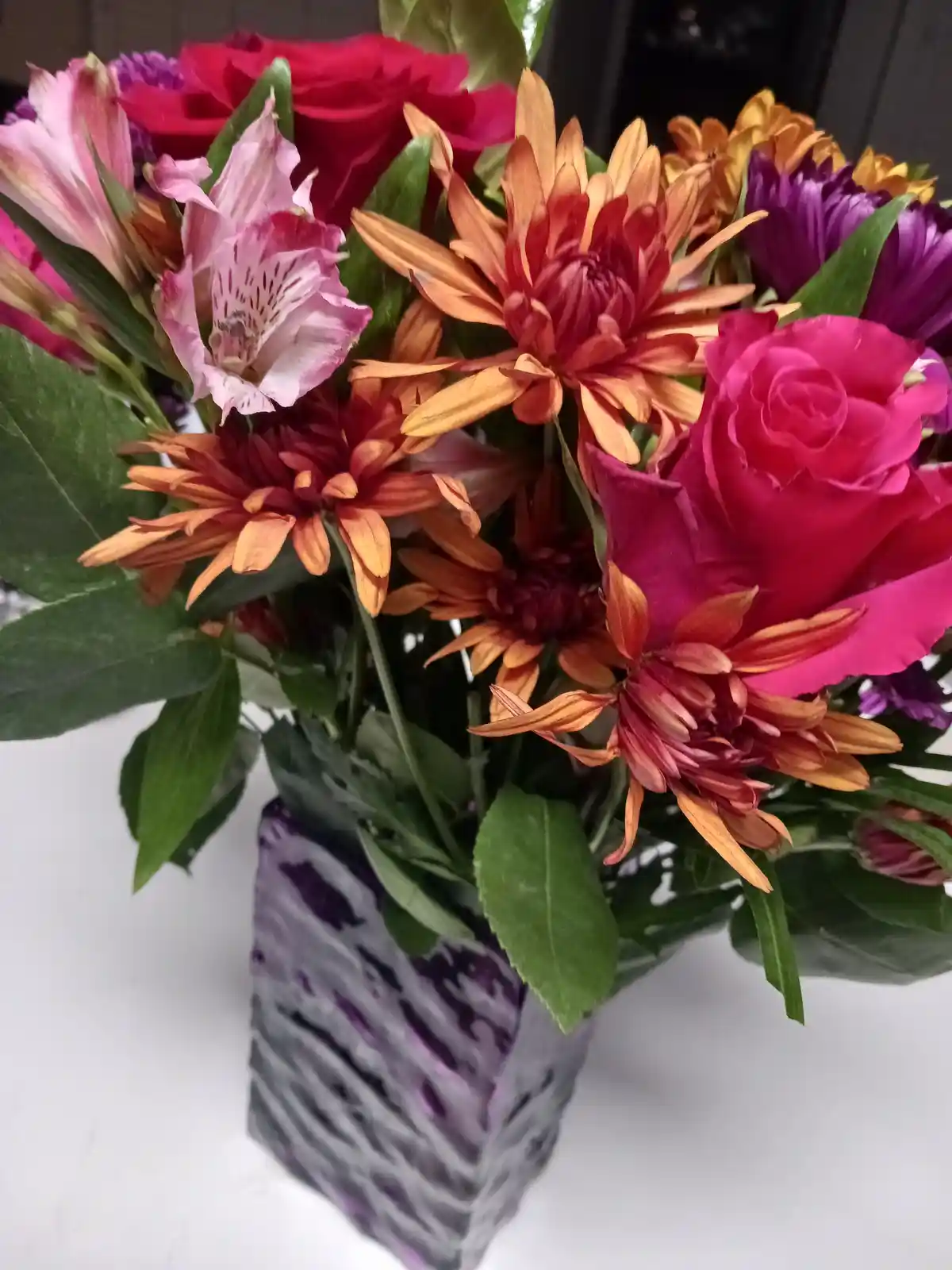 A vibrant bouquet of assorted flowers including pink roses, orange and red chrysanthemums, and purple blooms arranged in a…