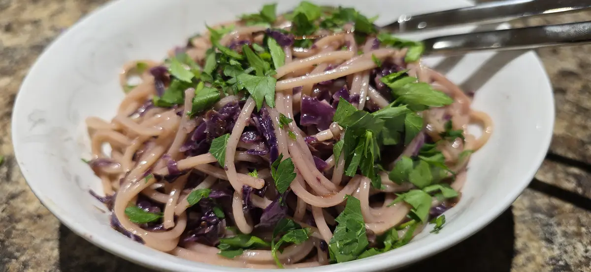 A close-up photo of a white bowl filled with light-colored noodles mixed with shredded purple cabbage and chopped green…