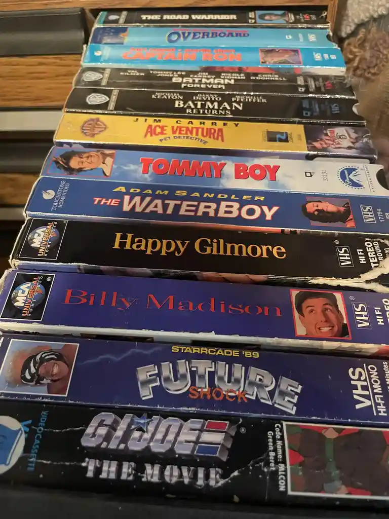 A stack of vintage VHS tapes featuring 1980s and 1990s movies like 'The Road Warrior,' 'Ace Ventura,' 'Tommy Boy,' 'The…