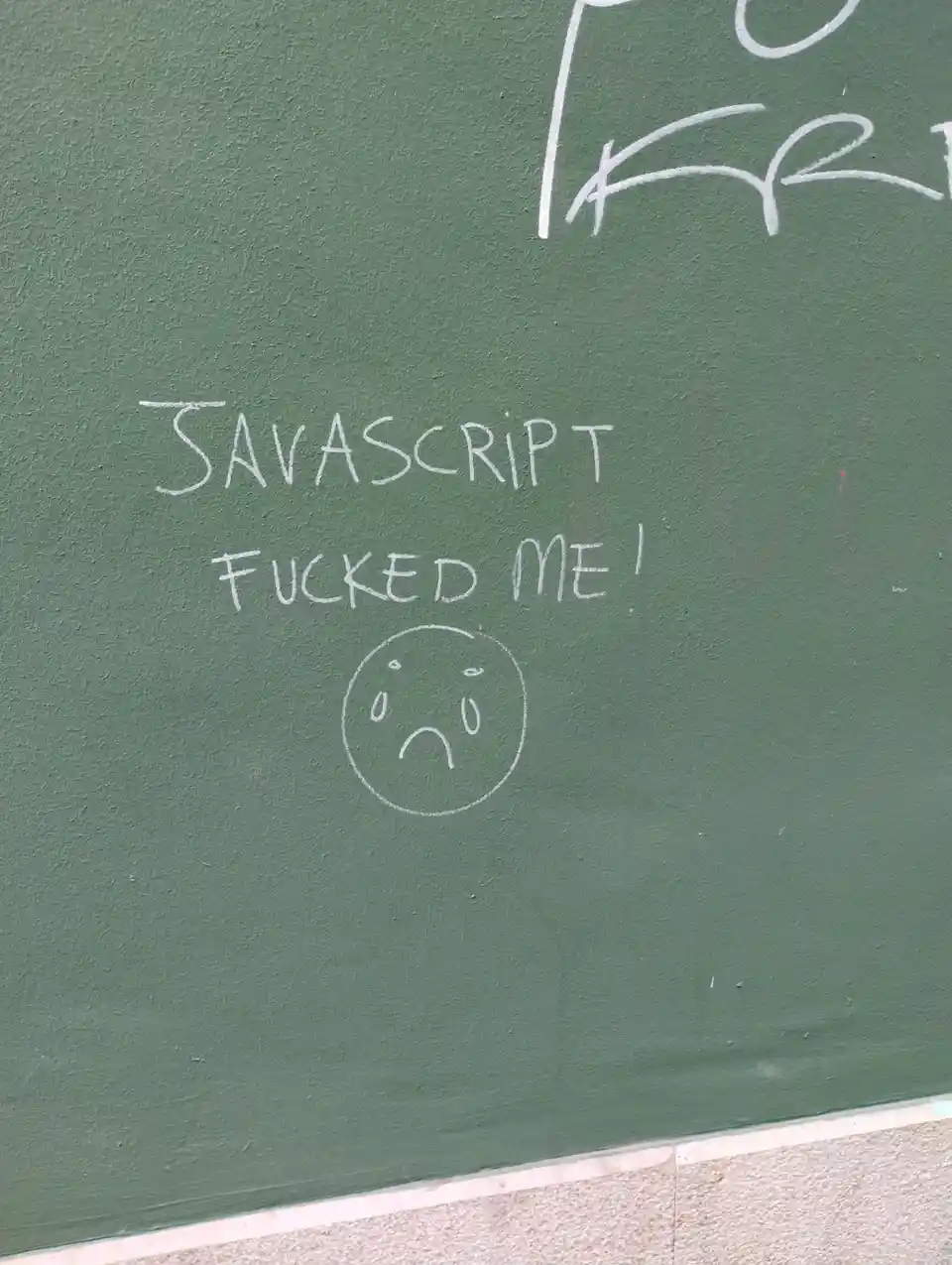 White chalk writing on a green chalkboard wall reads 'JAVASCRIPT FUCKED ME!' with a simple sad face drawn below. Partially…