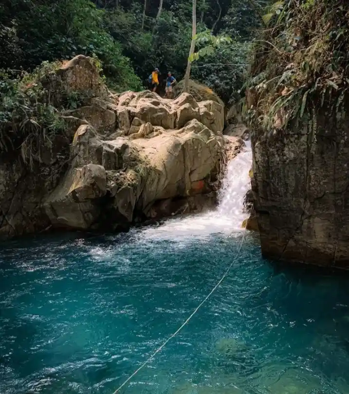 Two people stand on a rocky ledge above a turquoise river with a small waterfall. A thin rope stretches from the ledge down…