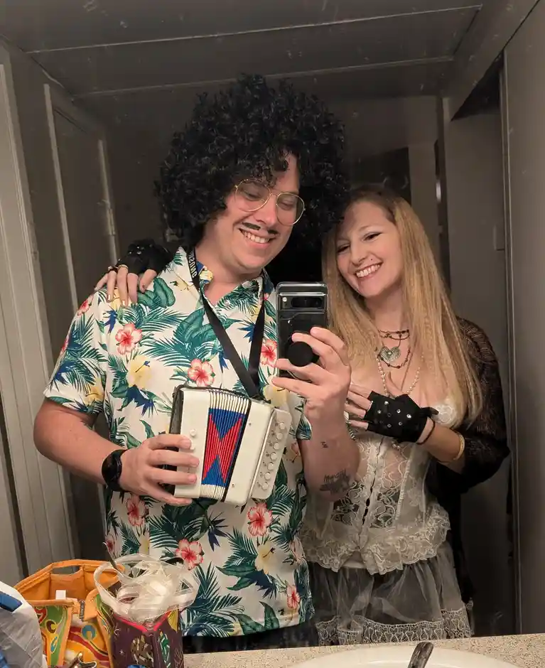 A man wearing a black curly wig, gold-rimmed glasses, and a mustache takes a selfie in a mirror while holding an accordion…
