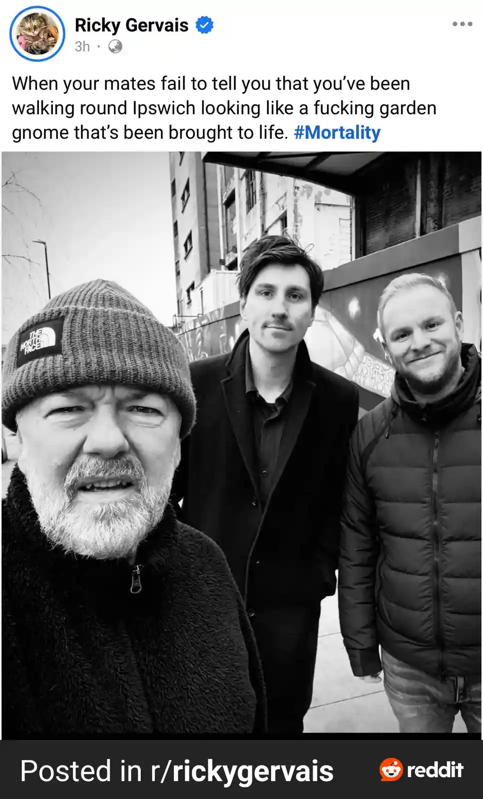 A black-and-white selfie shows Ricky Gervais in the foreground wearing a beanie and a fuzzy jacket, looking surprised or…
