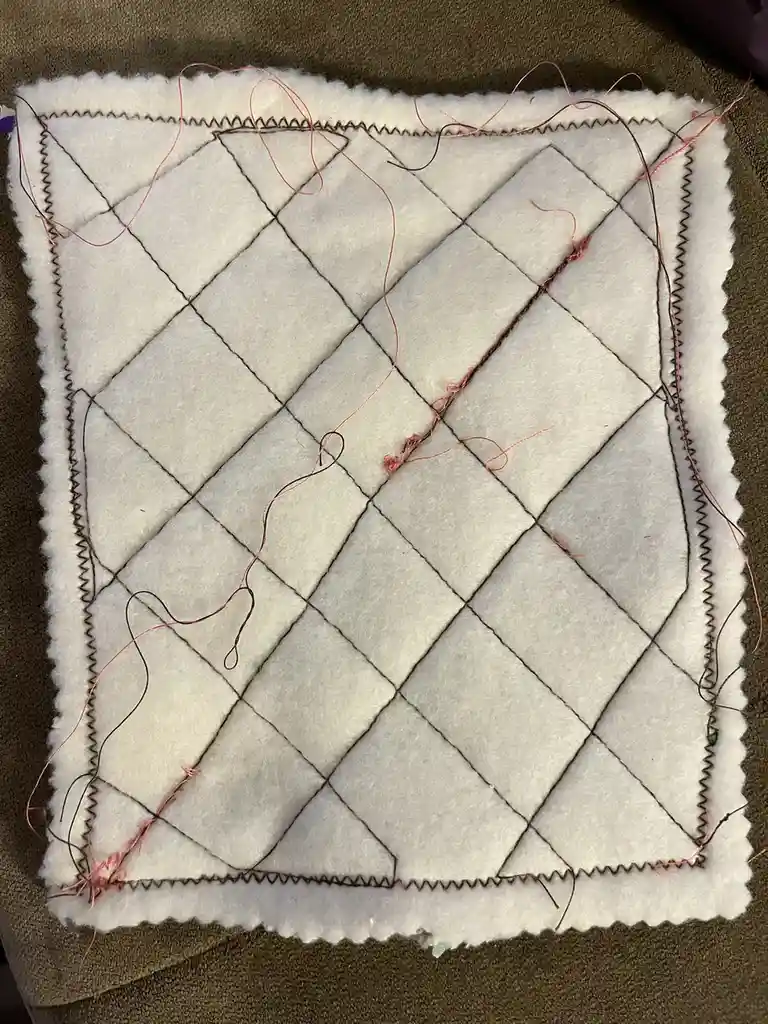 A handmade quilt square with a diamond pattern stitched in black thread on white felt, featuring loose red and black threads…