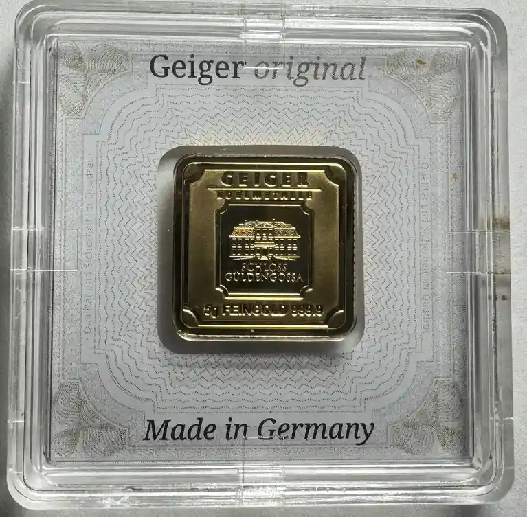 A square gold bar labeled 'GEIGER FEINMETALLE' with 'SCHLOSS GULDENGOSA' and '5g FEINGOLD 999.9' is displayed in a clear…
