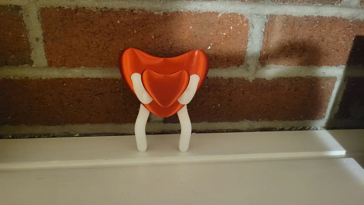 A small orange heart-shaped figurine with white arms and legs stands on a white windowsill in front of a brick wall, holding…
