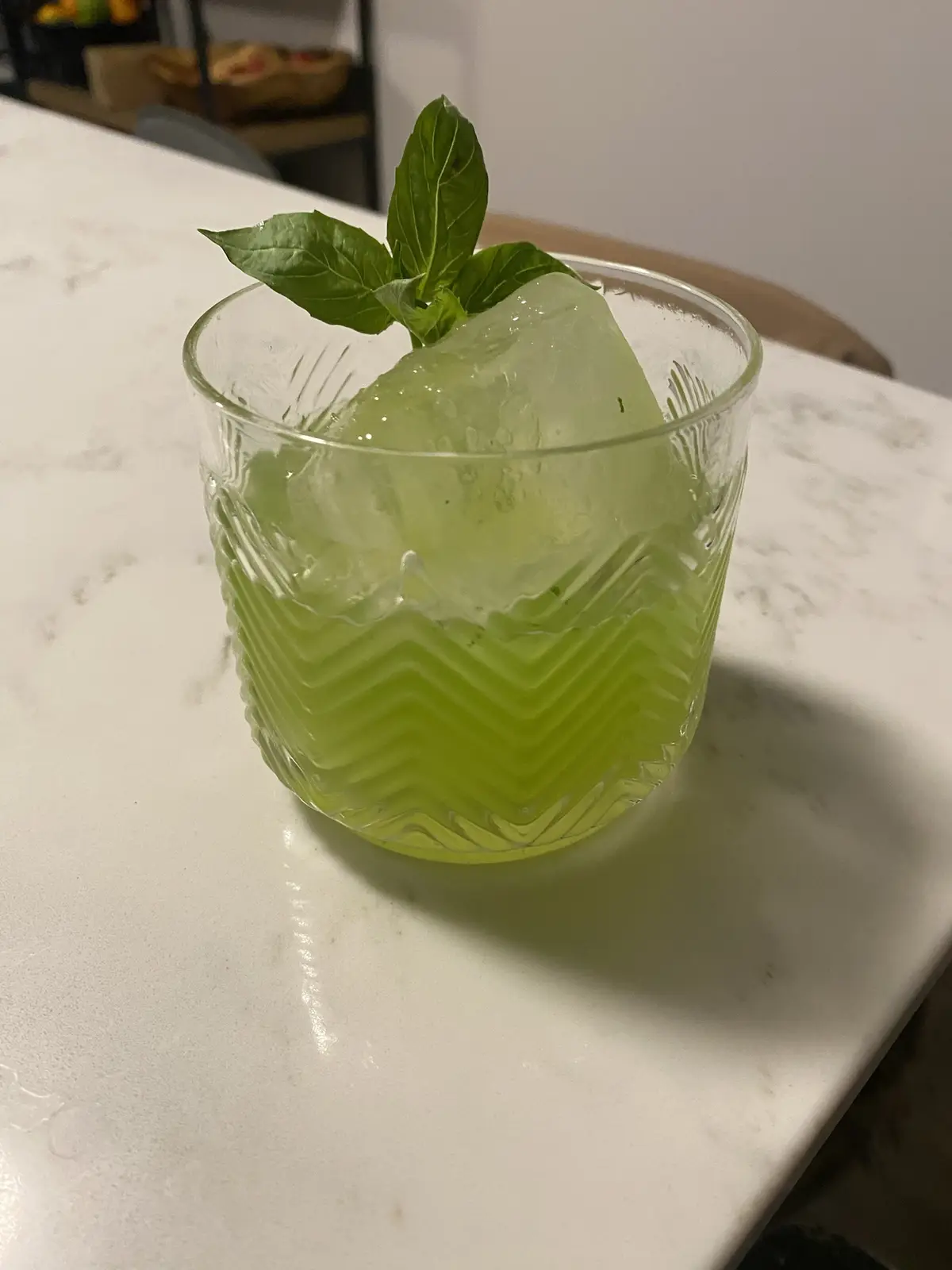 A close-up photo shows a glass with a large ice cube and a sprig of mint, filled with a pale green liquid, sitting on a…