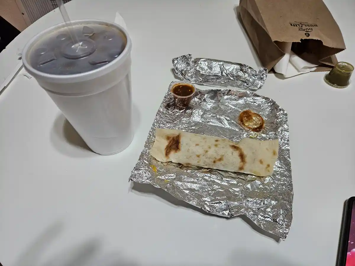 A white table holds a white styrofoam cup with a straw, a foil-wrapped burrito, a small cup of sauce, and a brown paper bag…
