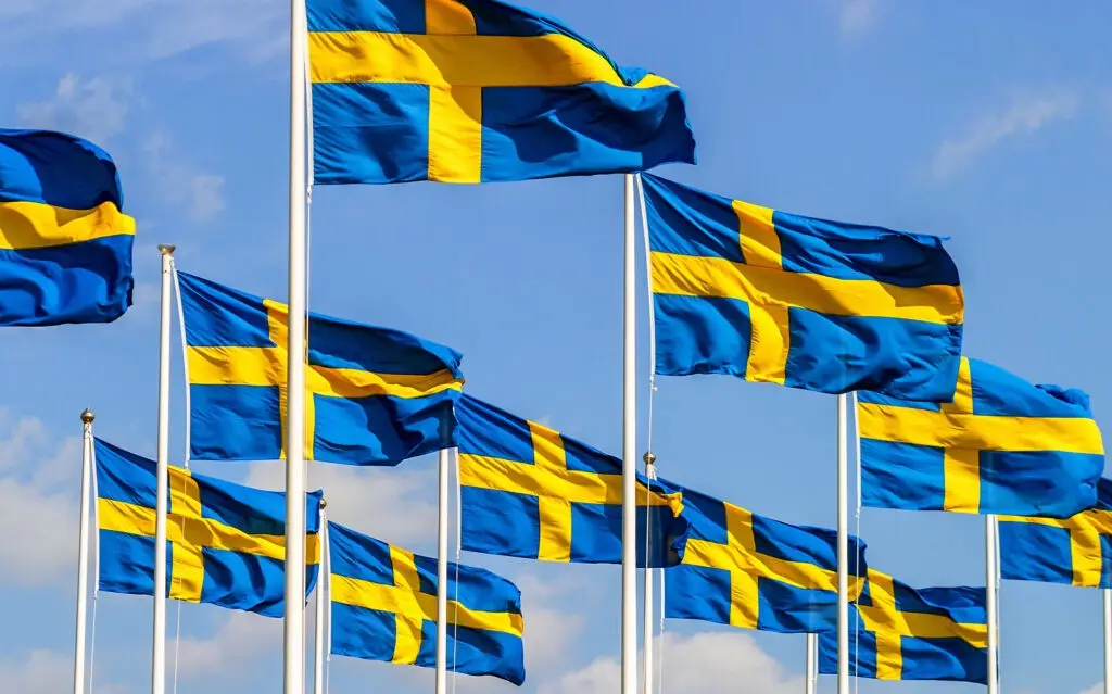 Multiple swedish flags with blue backgrounds and yellow crosses are waving on poles against a clear blue sky with scattered…