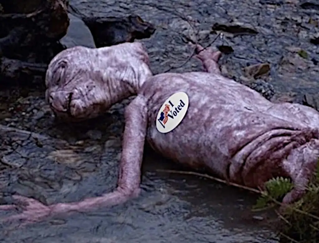 A pale, emaciated, wrinkled alien creature lies motionless on a rocky, muddy surface. A circular white sticker with the text…