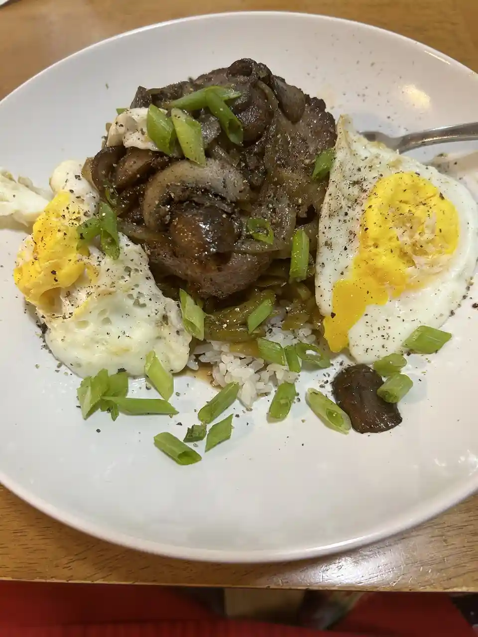 A close-up photo of a white bowl containing rice topped with cooked meat, mushrooms, and two fried eggs with runny yolks…