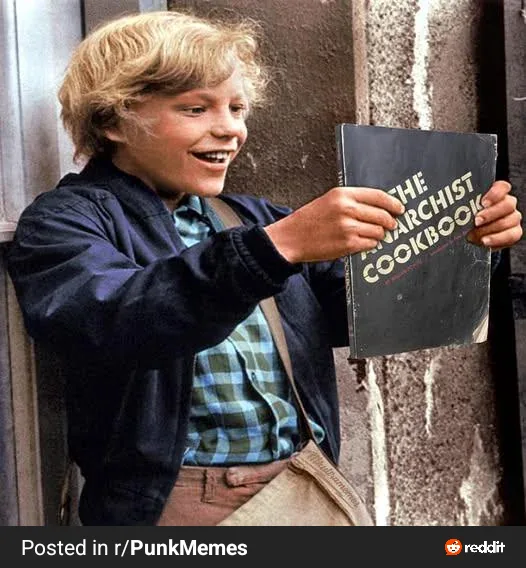 A young boy with blonde hair, wearing a dark jacket over a plaid shirt, smiles broadly while holding up a book titled 'THE…