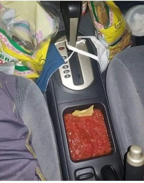 A car's center console has a cup holder filled with red salsa and a single tortilla chip, surrounded by bags of chips and a…