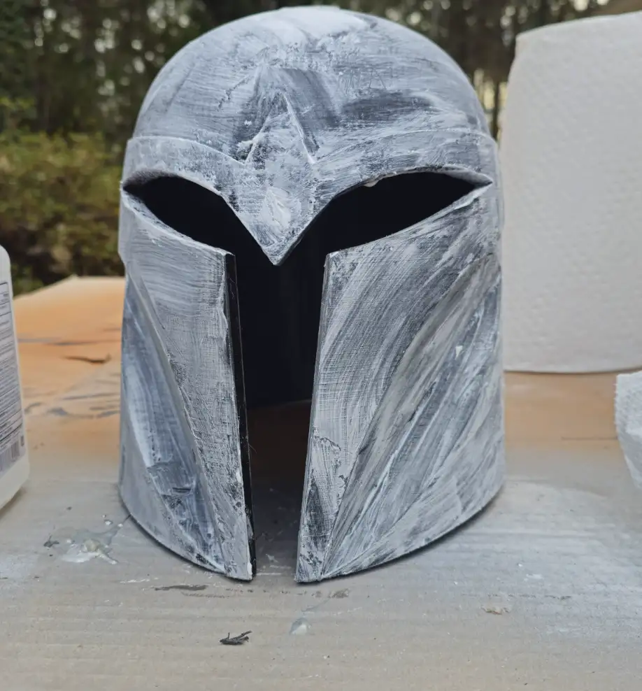 A handmade Mandalorian helmet, freshly painted with visible brush strokes and uneven gray paint, sits on a work surface…