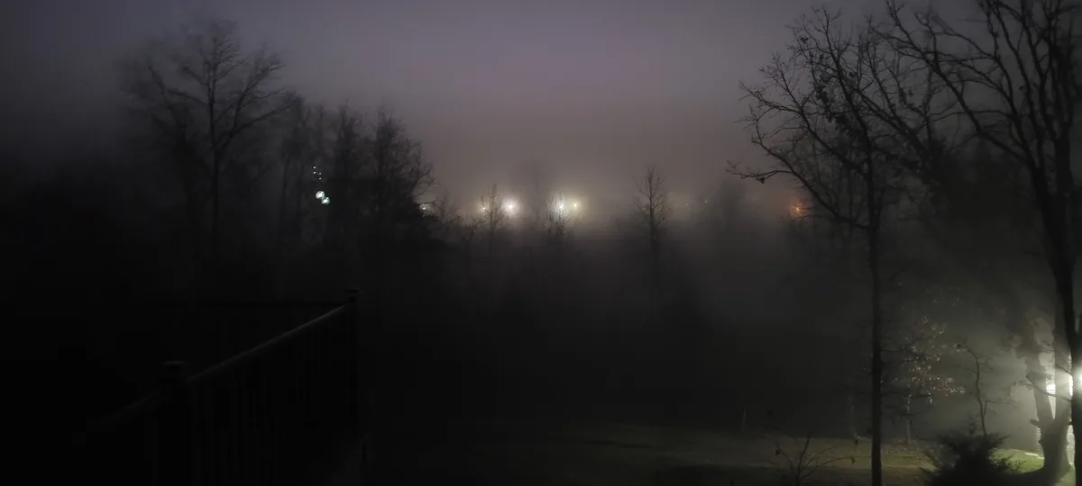 A dark, foggy night scene viewed from a deck, showing bare trees and distant lights obscured by mist. The overlaid text…
