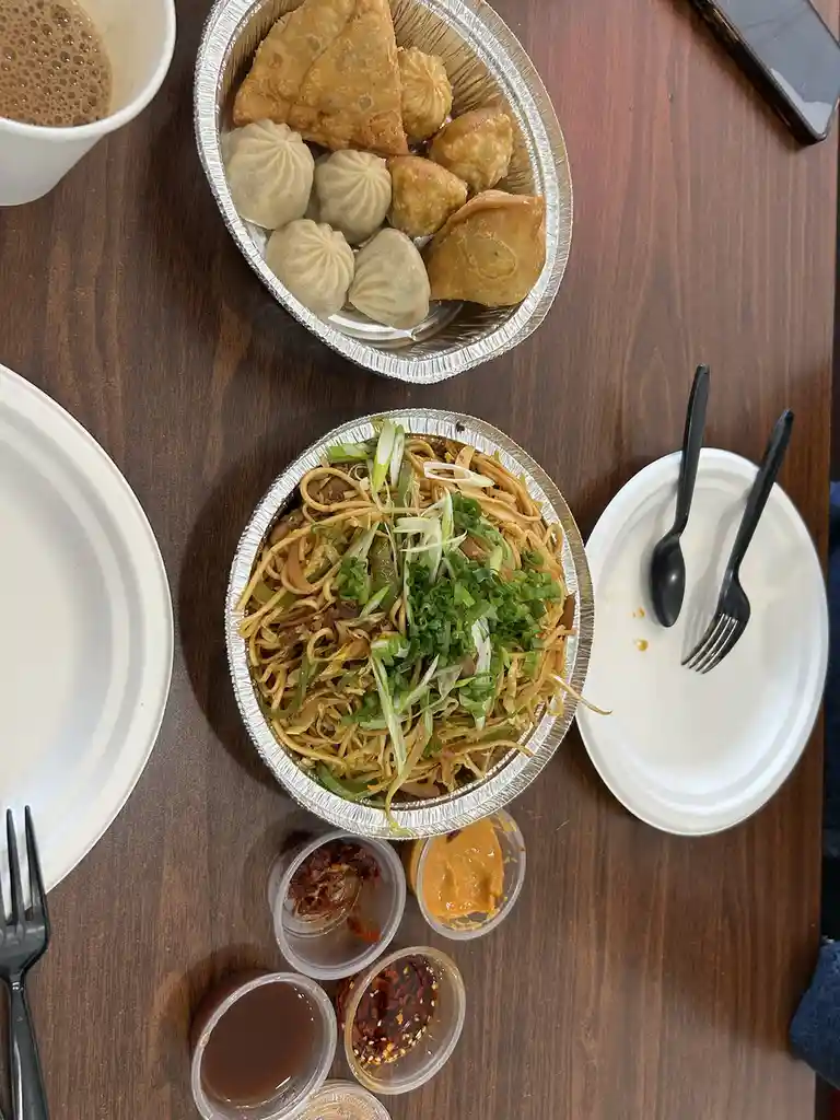 An overhead photo shows a wooden table with two foil containers: one holds fried samosas and dumplings, the other contains…