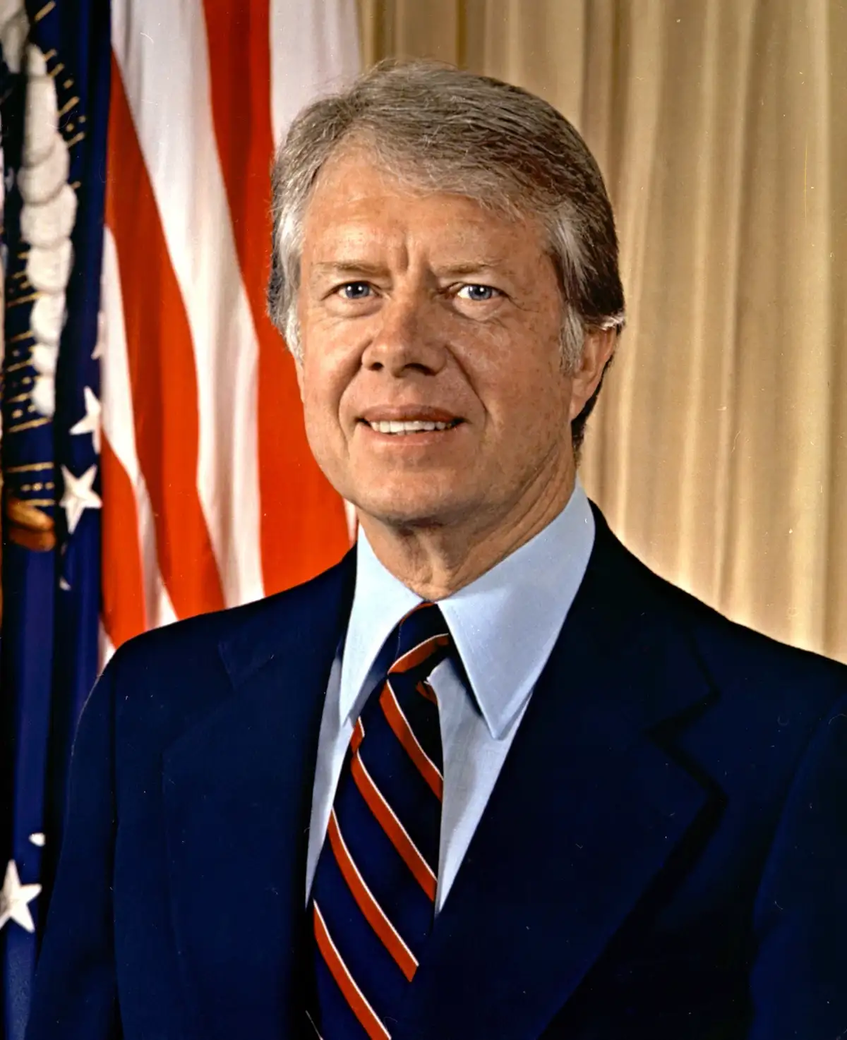 A formal portrait of Jimmy Carter, the 39th president of the United States, smiling while wearing a dark blue suit, light…