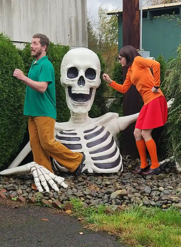 A man in a green shirt and brown pants stands beside a large skeleton statue, looking shocked as a woman in an orange top…