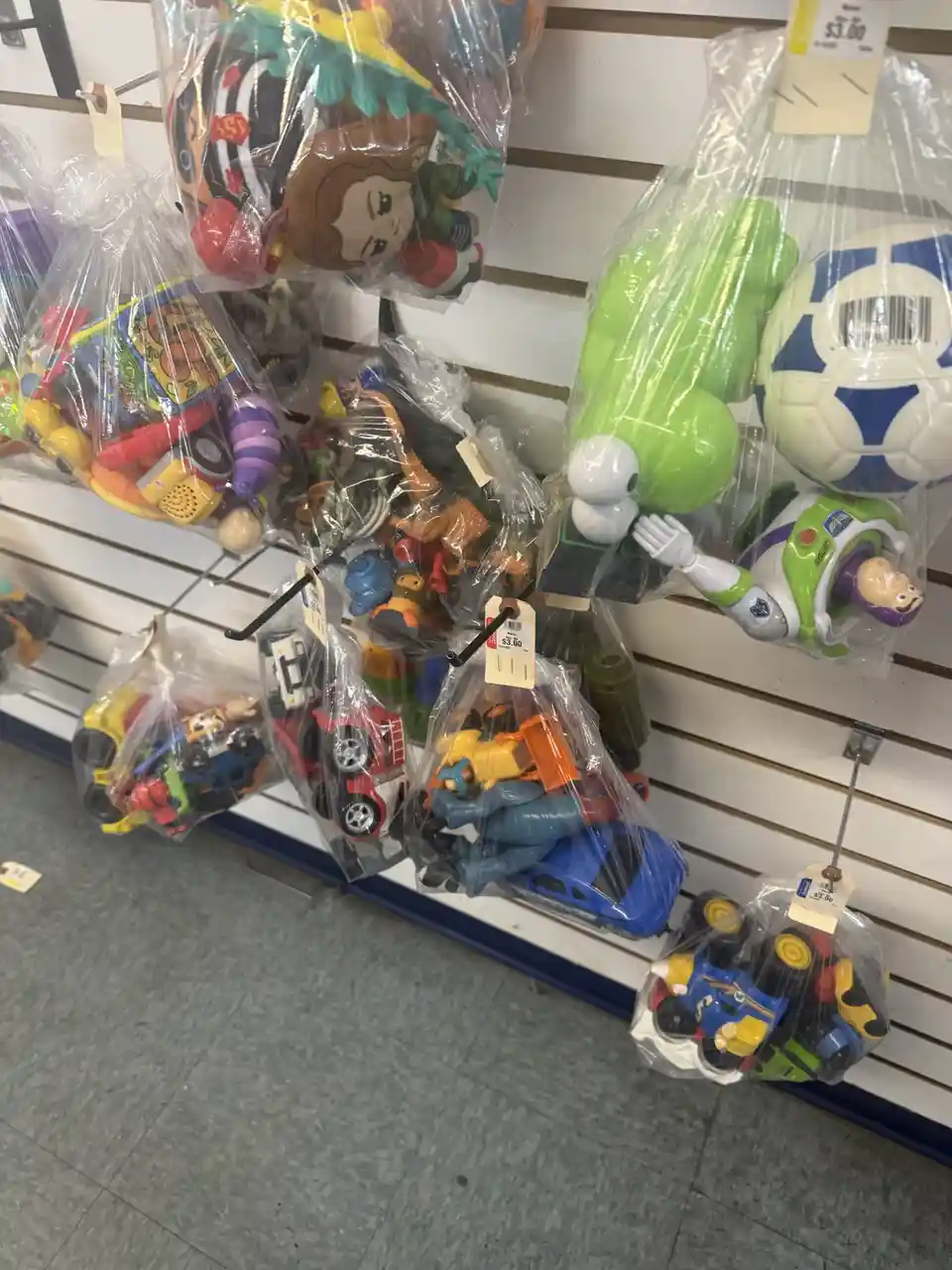 A store shelf displays numerous bags of assorted cheap toys, including Buzz Lightyear, plush dolls, and toy cars, all…
