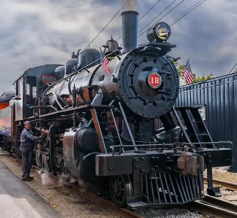 A man in overalls stands beside a large, black steam locomotive marked with the number 18 and American flags, as steam rises…