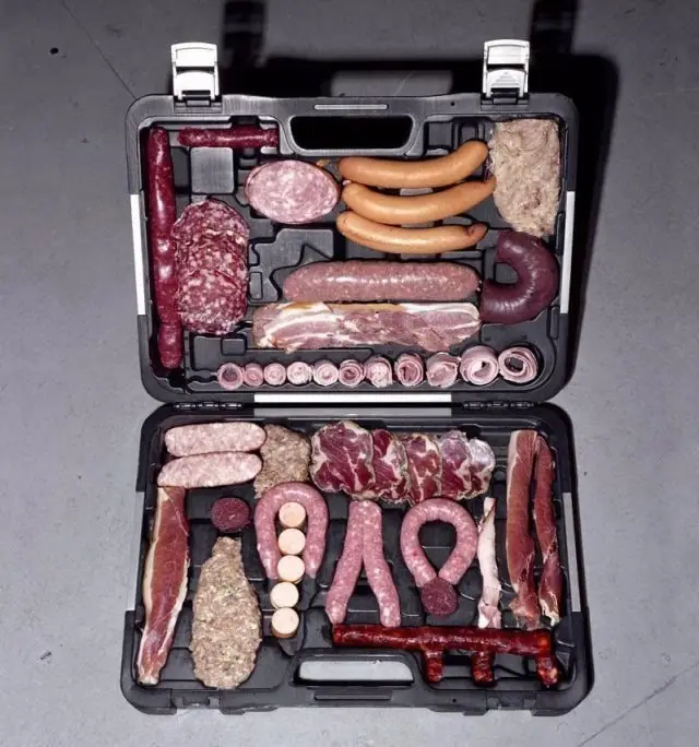 An open black plastic toolbox is filled with various types of raw meat, including sausages, salami, bacon, and other…