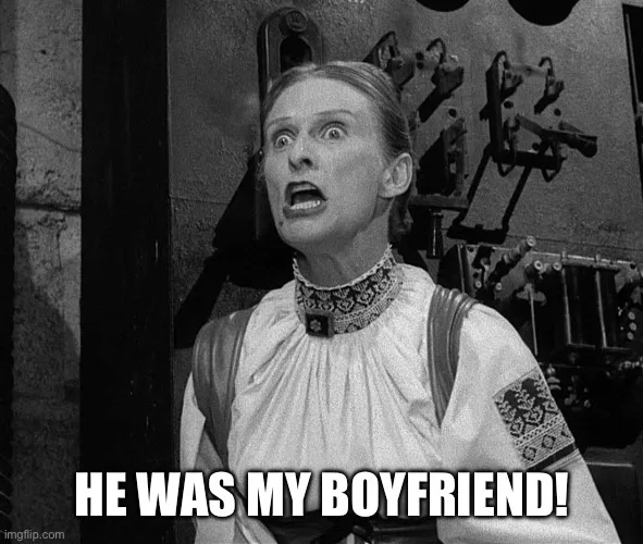 A black-and-white still of a woman with wide eyes and an open mouth, looking shocked or horrified. She wears a…