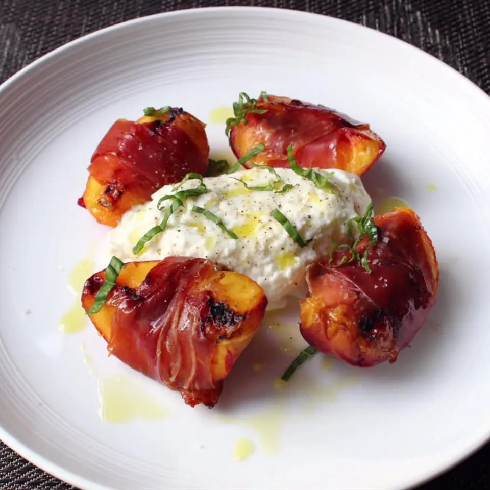 A white plate holds four grilled peach halves wrapped in crispy bacon, arranged around a central mound of creamy burrata…