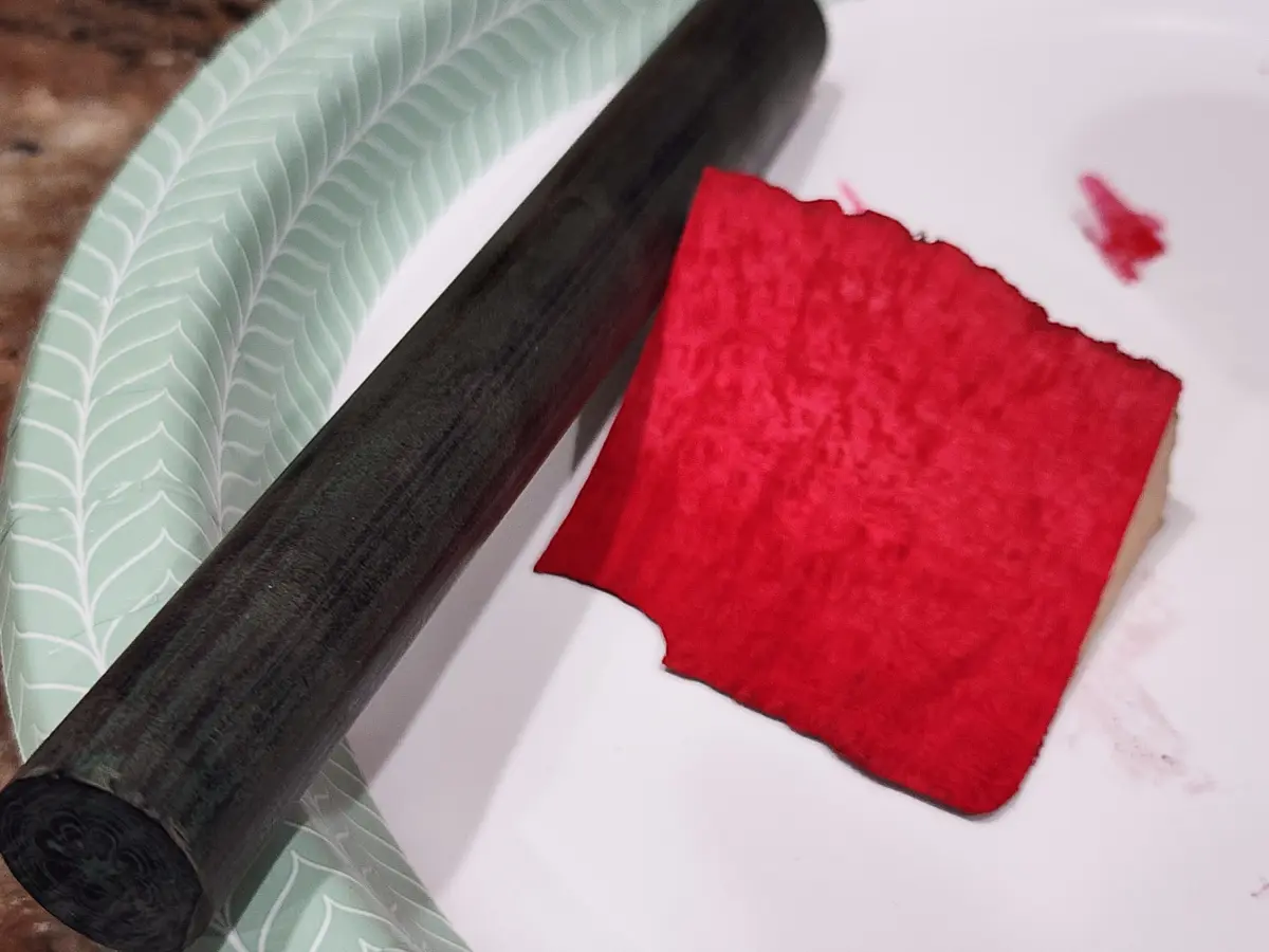 A dark wooden rolling pin rests on a light green plate with white leaf patterns, next to a square of vibrant red velvet cake…