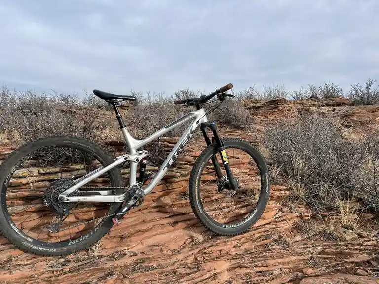 A silver Trek mountain bike is parked on reddish-orange rocky terrain with dry brush under an overcast sky. The bike has…