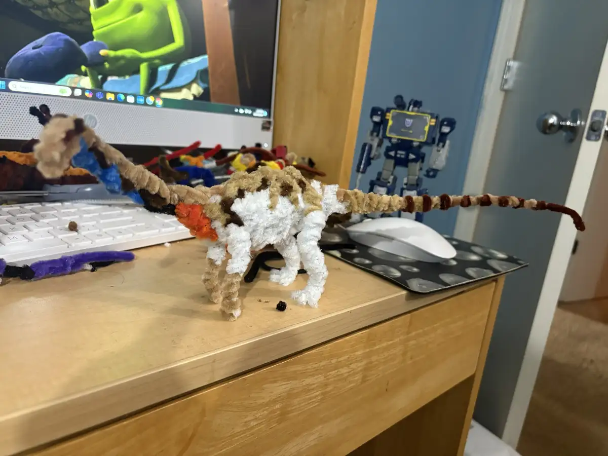 A handmade, fuzzy dinosaur crafted from pipe cleaners sits on a wooden desk next to a white keyboard and mouse. In the…