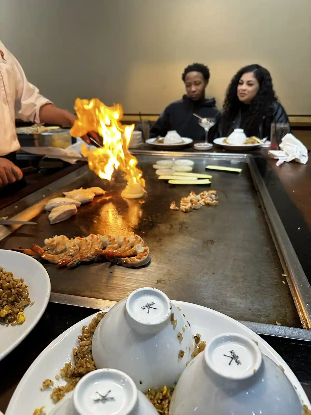 A teppanyaki restaurant scene where a chef is flambeing food on a large grill, with flames erupting from a piece of onion…