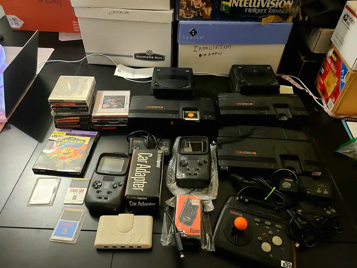 A cluttered tabletop displays a collection of vintage NEC TurboGrafx-16 hardware, including multiple consoles, controllers…