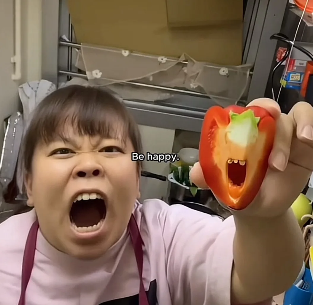 A woman in a kitchen wearing an apron is screaming with her mouth wide open while holding up a red bell pepper that has been…