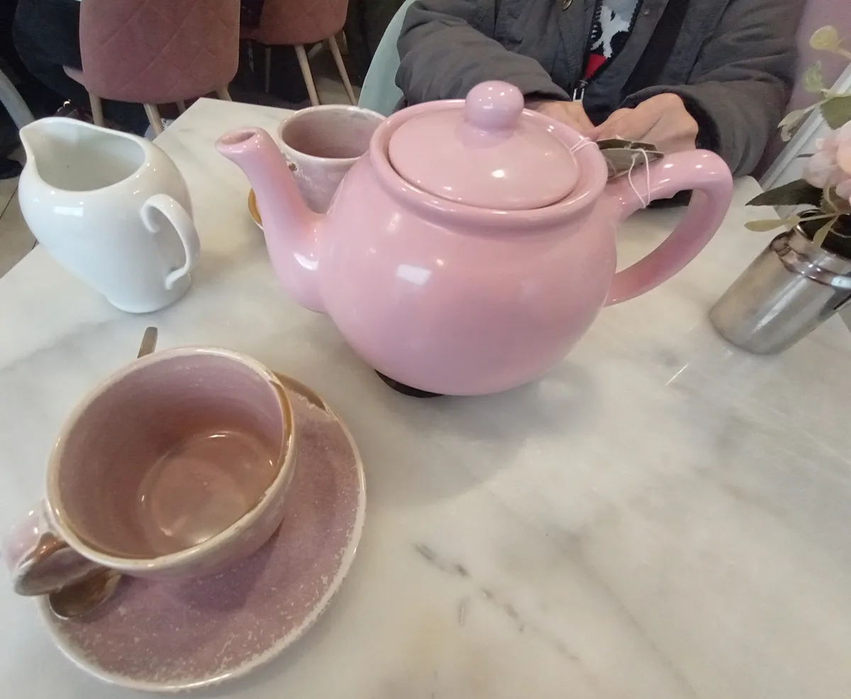 A close-up photo shows a table with a large pink teapot, a white creamer, and two pink ceramic cups on a marble surface. One…